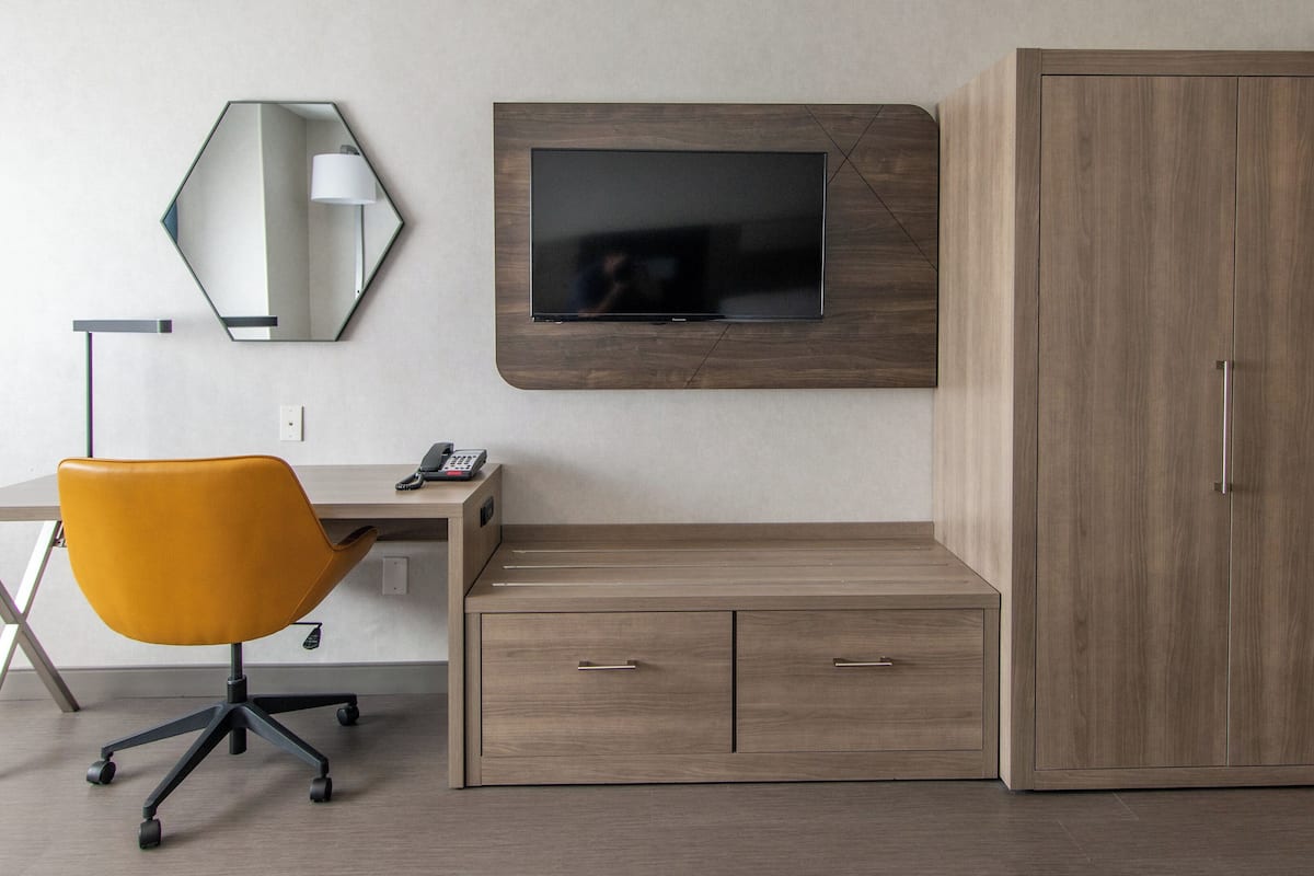 Una habitación de hotel moderna con un escritorio, una silla de oficina amarilla, un armario grande, un televisor montado en la pared y un espejo adosado.