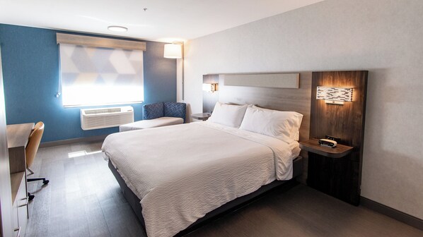 Une chambre d’hôtel moderne avec un grand lit, un bureau et une chaise. On y trouve une fenêtre avec des stores, un climatiseur fixé au mur et une table de chevet avec une lampe.