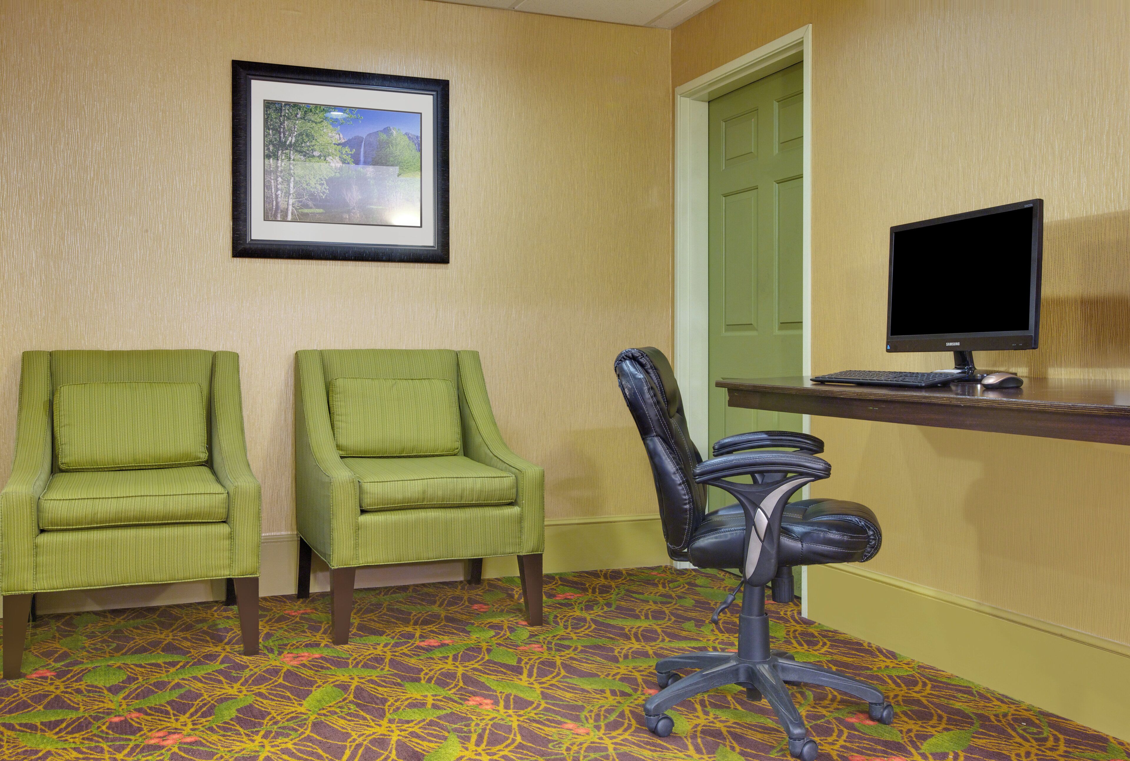 A room with two green armchairs, a black office chair, a desk with a computer monitor, and a framed picture on the wall.