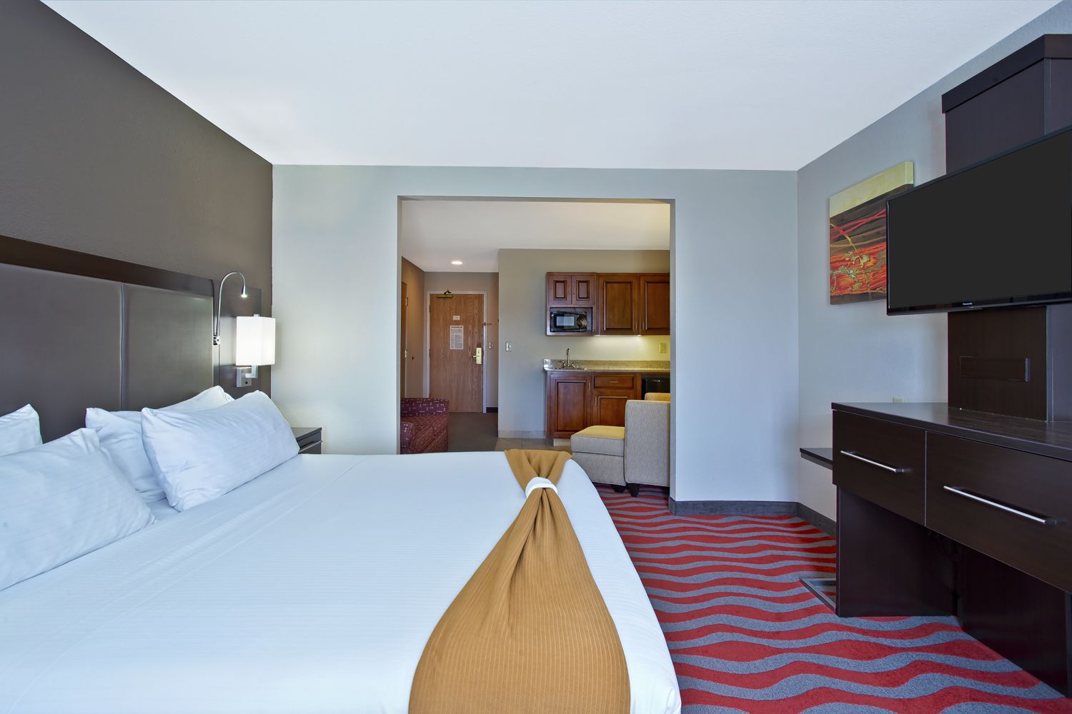A hotel room with a large bed, a TV, a desk, and a kitchenette.