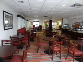 A hotel dining area with multiple tables and chairs, a red sofa, and a kitchen area in the background.