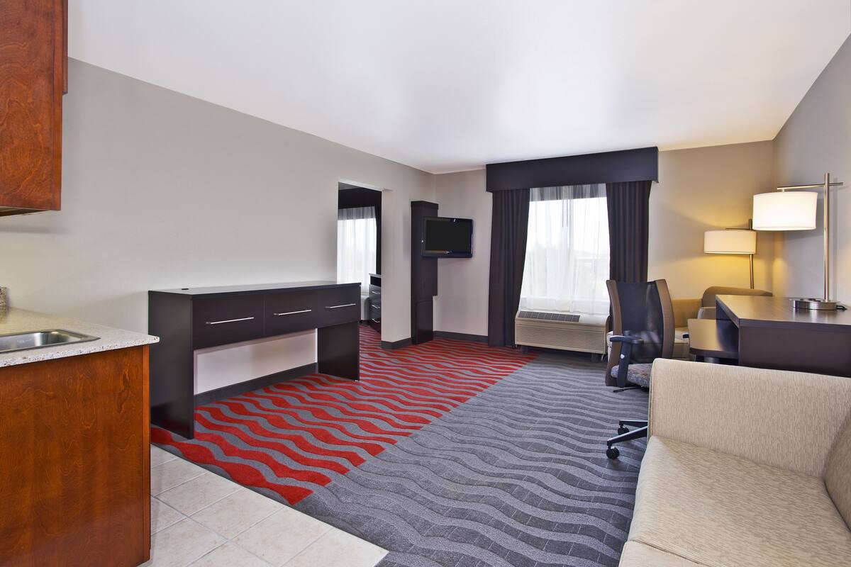 A hotel room with a kitchenette, a flat-screen TV mounted on the wall, a desk with a chair, and a sofa.