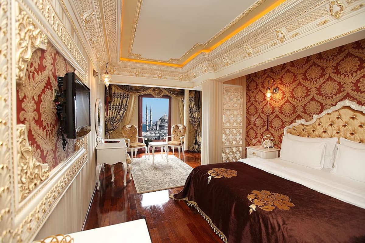 Une chambre d’hôtel luxueuse, avec un papier peint orné aux motifs dorés et rouges, un grand lit recouvert d’une courtepointe marron foncé, et une vue sur le paysage urbain par la fenêtre.