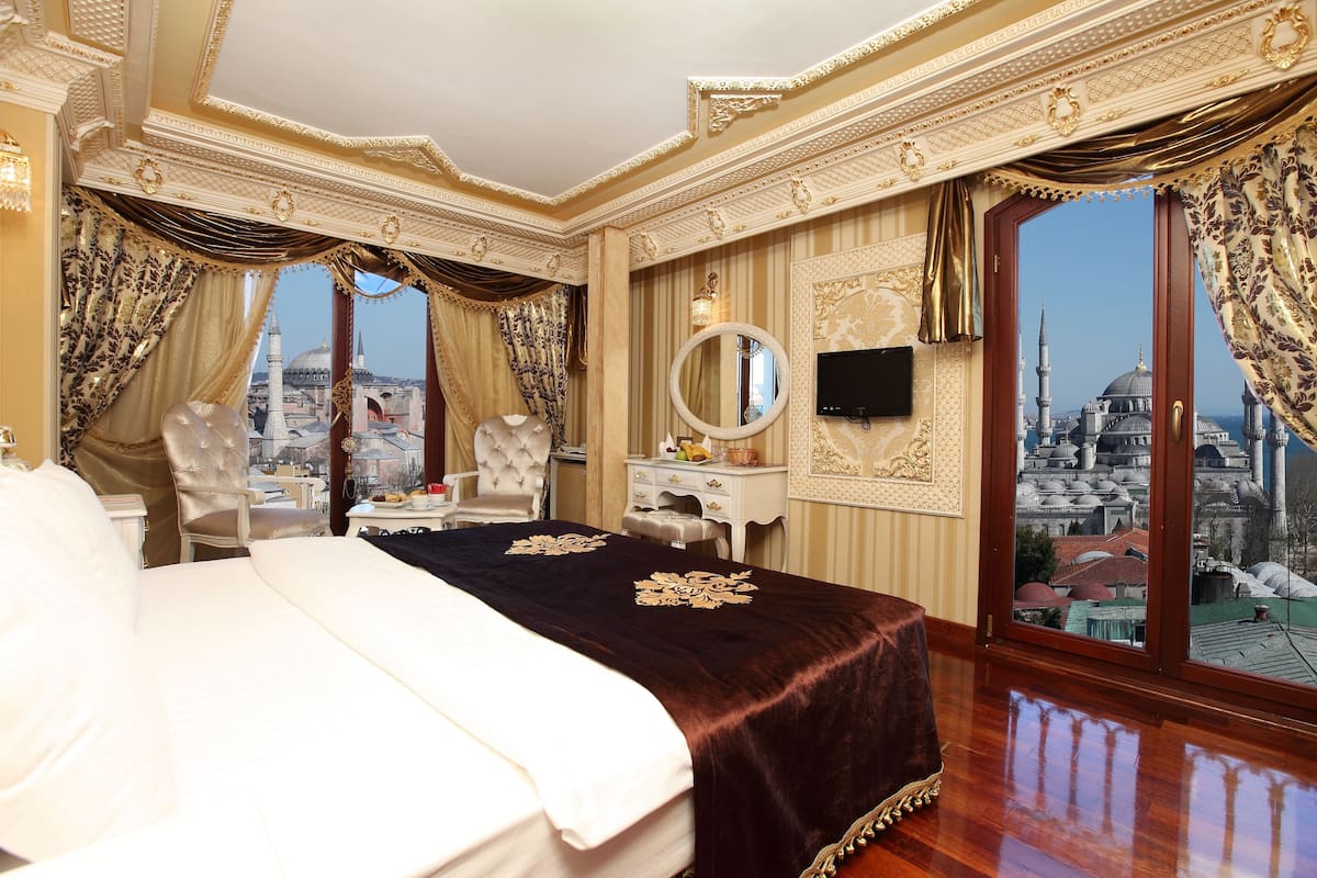 Une chambre d’hôtel luxueuse, dotée d’un grand lit, d’un coin salon avec un fauteuil et une table, d’une coiffeuse avec un miroir et offrant une vue sur le paysage urbain grâce à de grandes fenêtres.