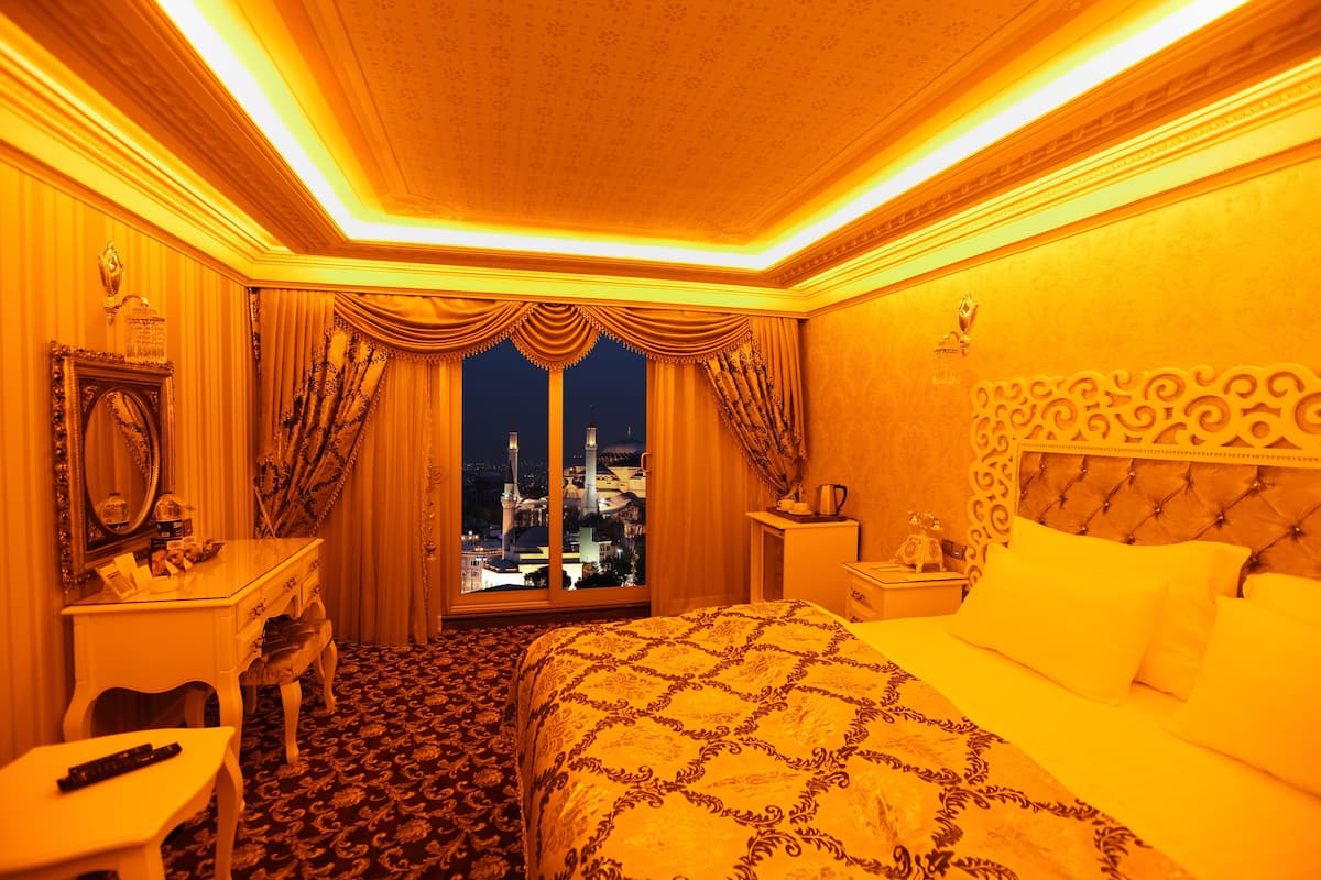 Une chambre d’hôtel avec un lit, une table de chevet, une commode, un miroir et une fenêtre donnant sur une vue panoramique de la ville la nuit.