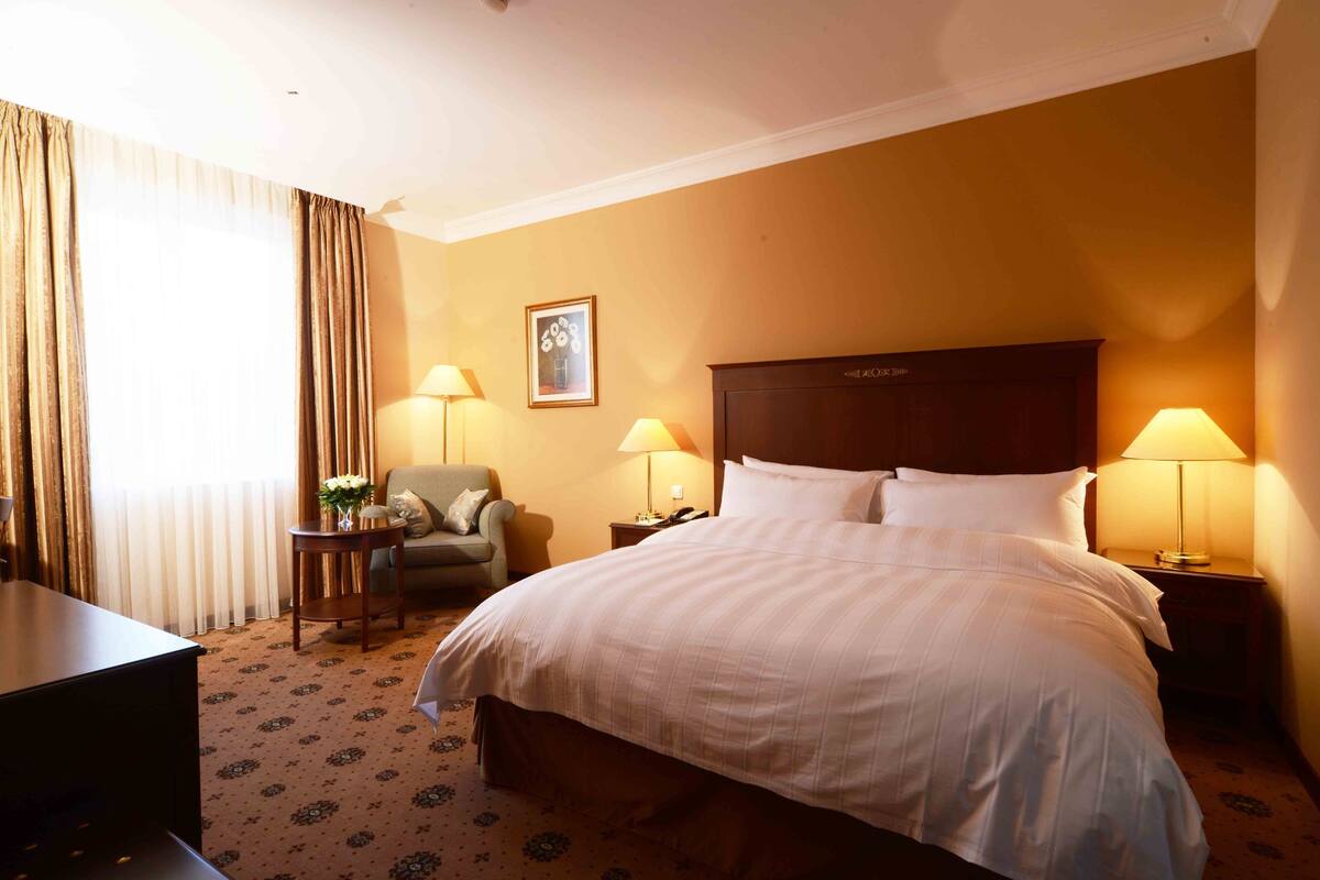 Habitación de hotel con una cama grande, dos mesitas de noche con lámparas, una silla, un escritorio y un cuadro en la pared.