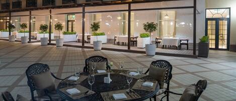 An outdoor dining area with round tables and chairs, surrounded by potted plants and a building with multiple balconies.