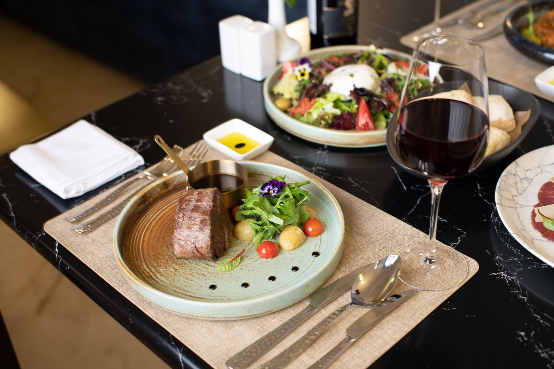 Una mesa de comedor con platos de comida, una copa de vino tinto y cubiertos.