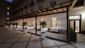 Un espacio al aire libre cubierto con mesas y sillas, plantas en macetas y un edificio con varias ventanas y balcones.