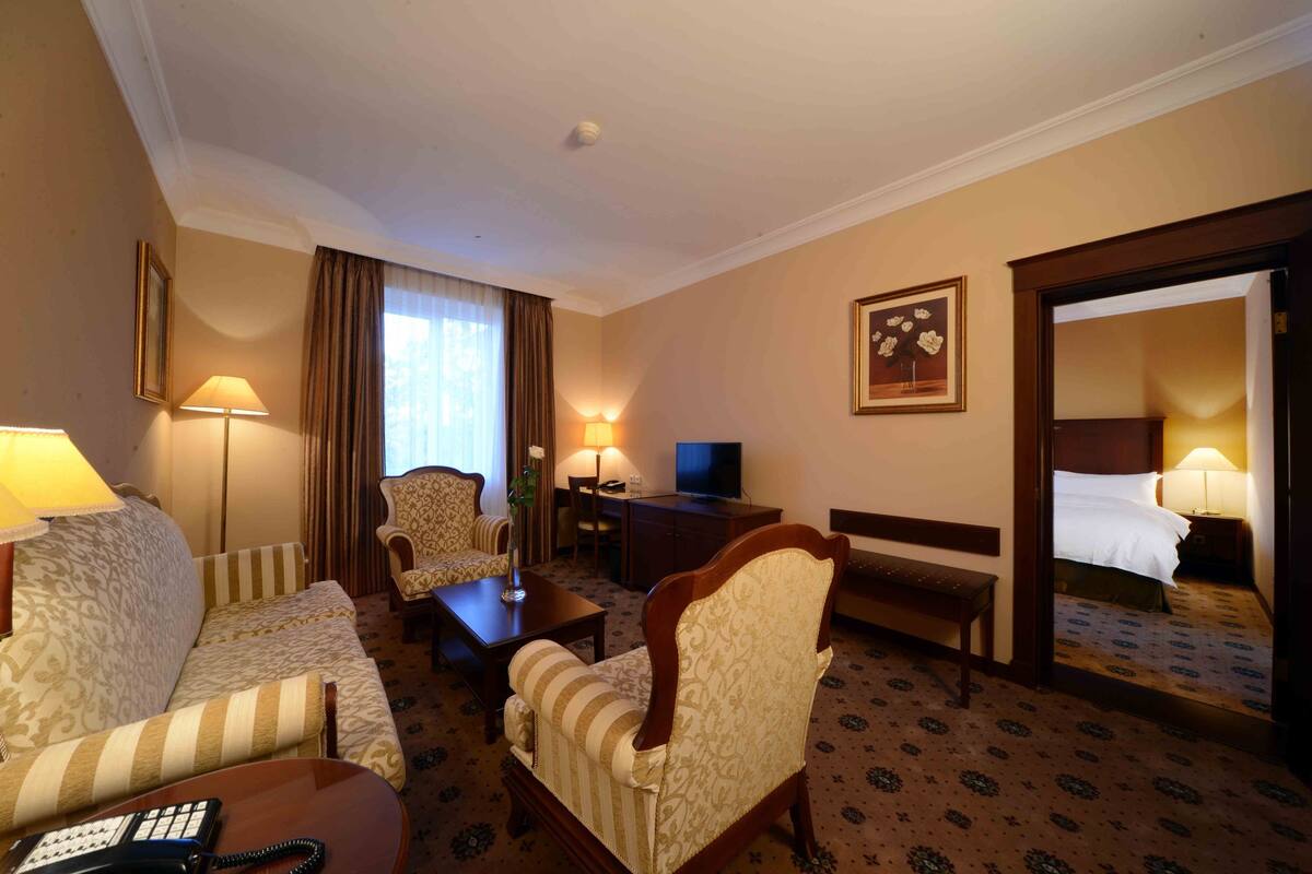Una habitación de hotel con sofá, sillón, una mesita, escritorio con televisor, mesita de noche con lámpara y cama.