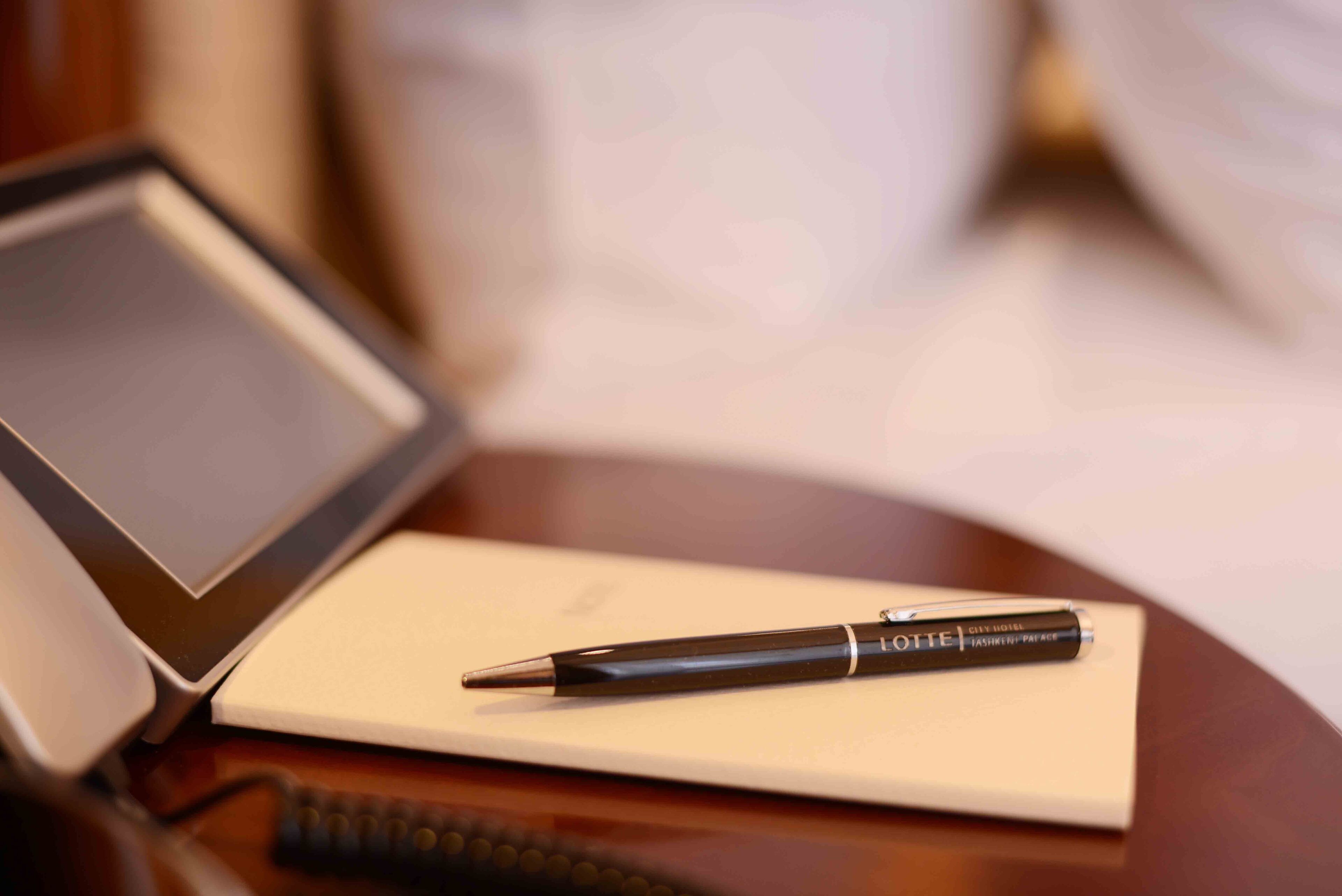 A closed laptop, a black pen on a notepad, and a telephone on a wooden surface.