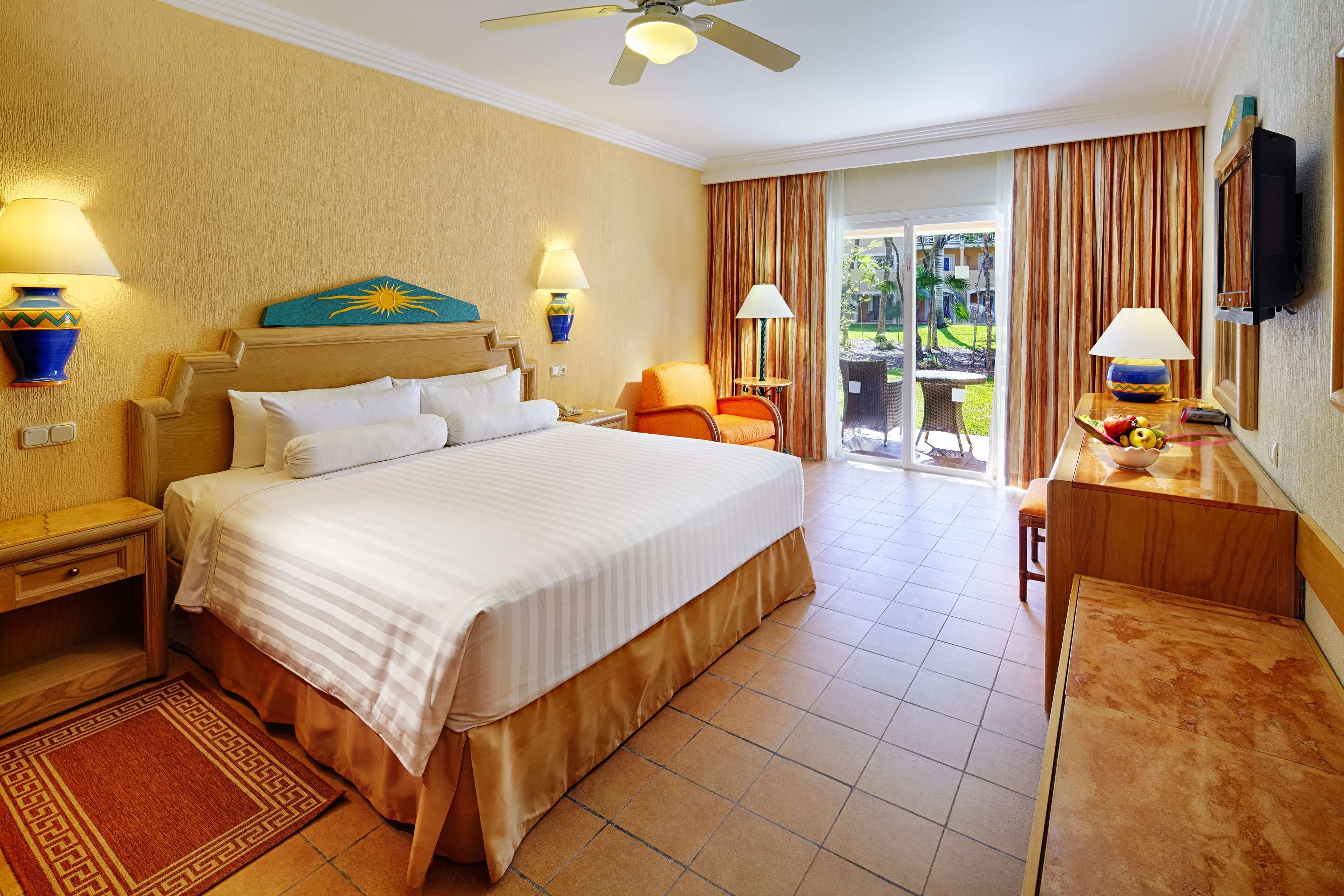A hotel room with a large bed, two bedside tables with lamps, a desk with a chair, and a view of the outdoors through a sliding door.