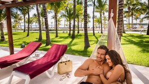 Una pareja descansando en una hamaca con vista a la playa, dos sillas de descanso con cojines rojos y un bolso de paja en el suelo.