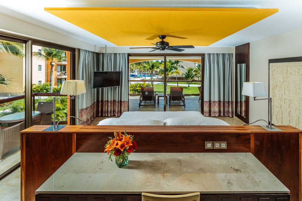 A hotel room with a bed, a desk, a chair, a TV, a ceiling fan, and a view of the outdoors.