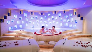 Una pareja en un jacuzzi circular rodeado de pétalos de rosa, con un panel de luces iluminadas al fondo.