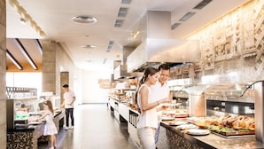 Zona de bufé con diversas opciones de comida, una cocina moderna al fondo y piso de baldosas.