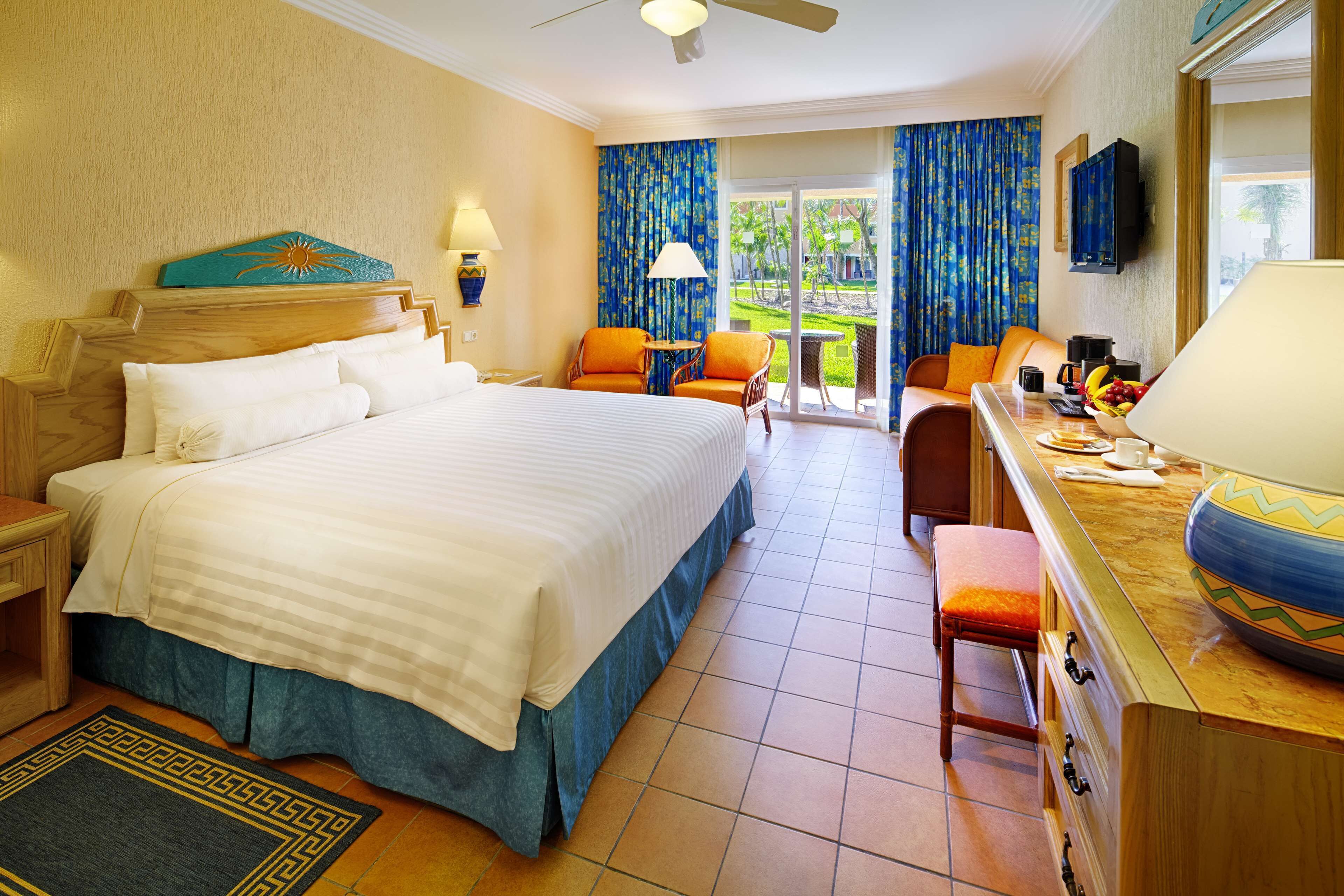 A hotel room with a large bed, a desk with a chair, a small table, and a view of the outdoors through a sliding door.