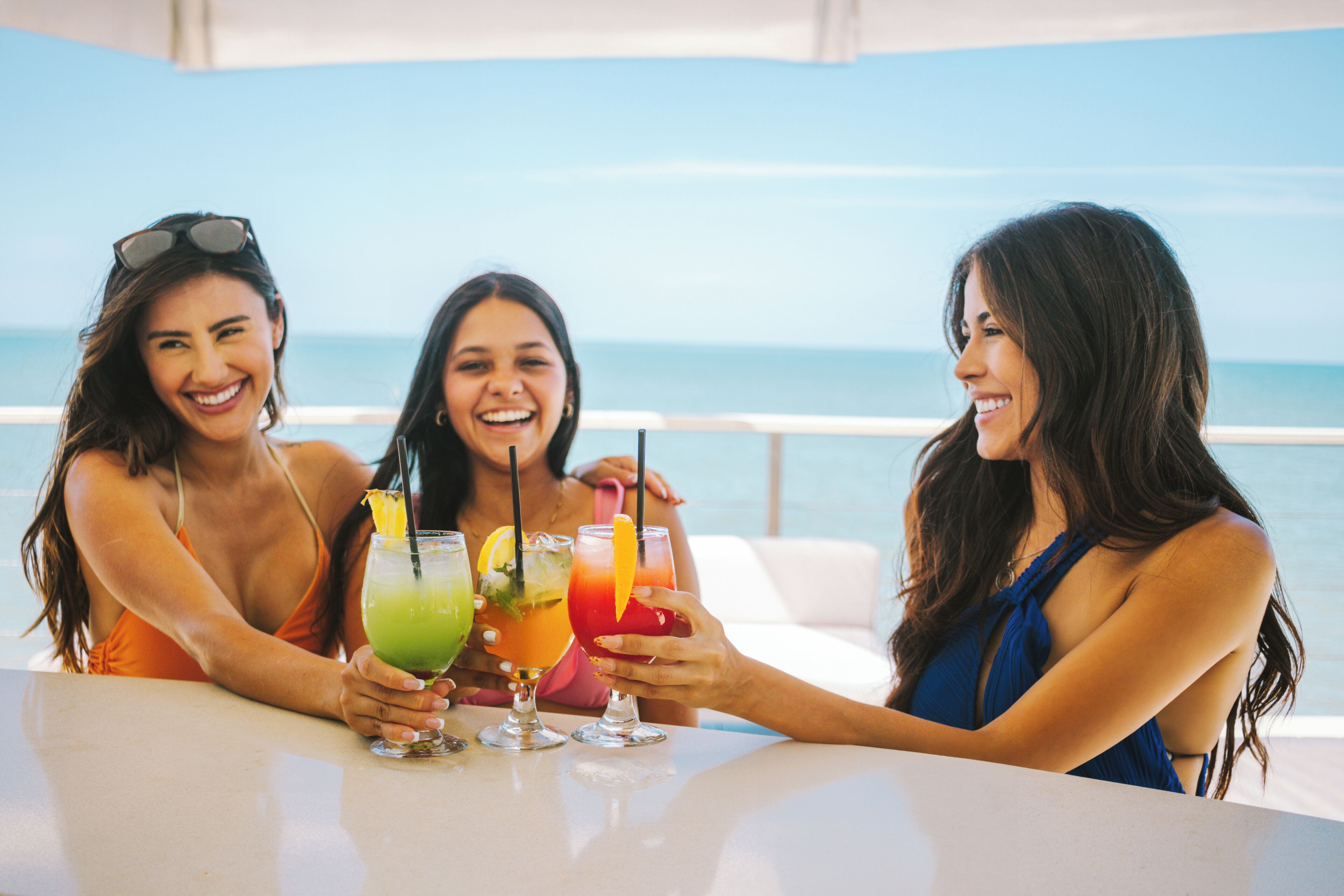 Trois femmes sont assises à une table, devant elles, des boissons, et elles ont une vue sur une étendue d’eau.