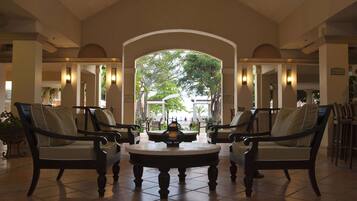 A lobby with a central round table, two armchairs, and a view through an arched doorway to an outdoor area with trees and a building.