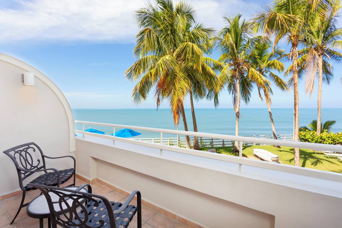 A balcony with two chairs, a view of the ocean, and palm trees.