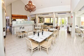 A spacious dining area with multiple tables and chairs, a central bar area, and large windows providing natural light.