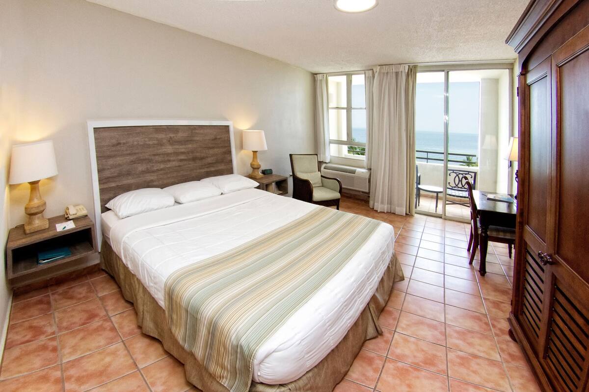 A hotel room with a large bed, a desk, a chair, and a view of the sea.