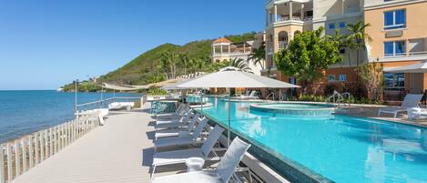 A poolside area with lounge chairs and umbrellas, a clear view of the sea, and a building with balconies.