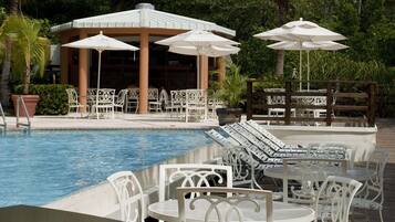 A pool area with white metal chairs and tables, umbrellas, and a covered seating area with a thatched roof.