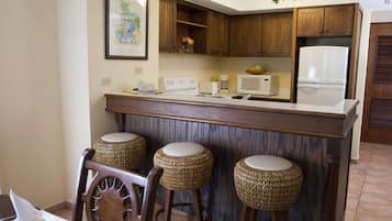 Una cocina con armarios de madera, un refrigerador blanco, un microondas y una cafetera. Hay tres taburetes de mimbre y una silla de madera.