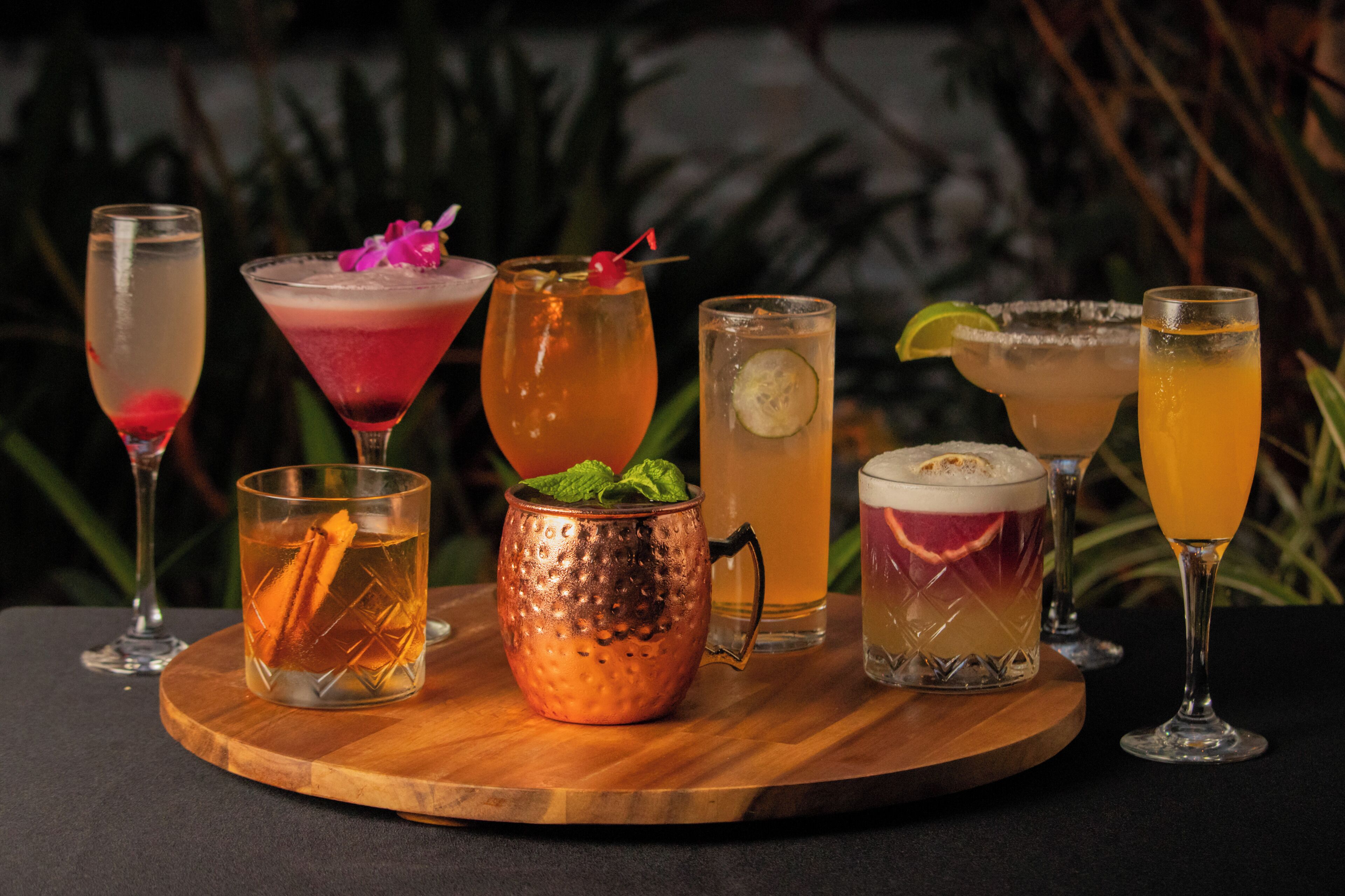 Une variété de boissons présentées sur une planche en bois, notamment un verre en cuivre, un cocktail margarita et un autre cocktail décoré d’une tranche de citron vert.