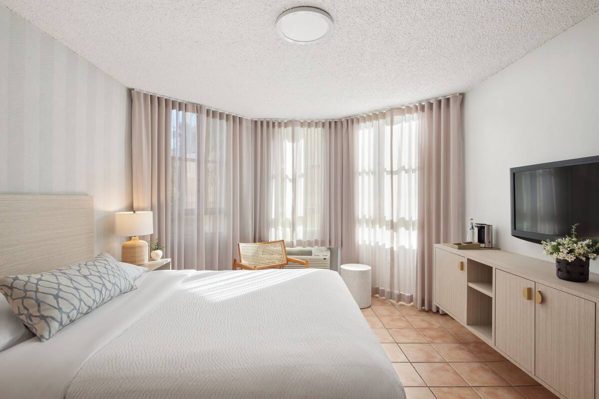 A modern hotel room with a large bed, a flat-screen TV, a desk with a lamp, and a large window with curtains.