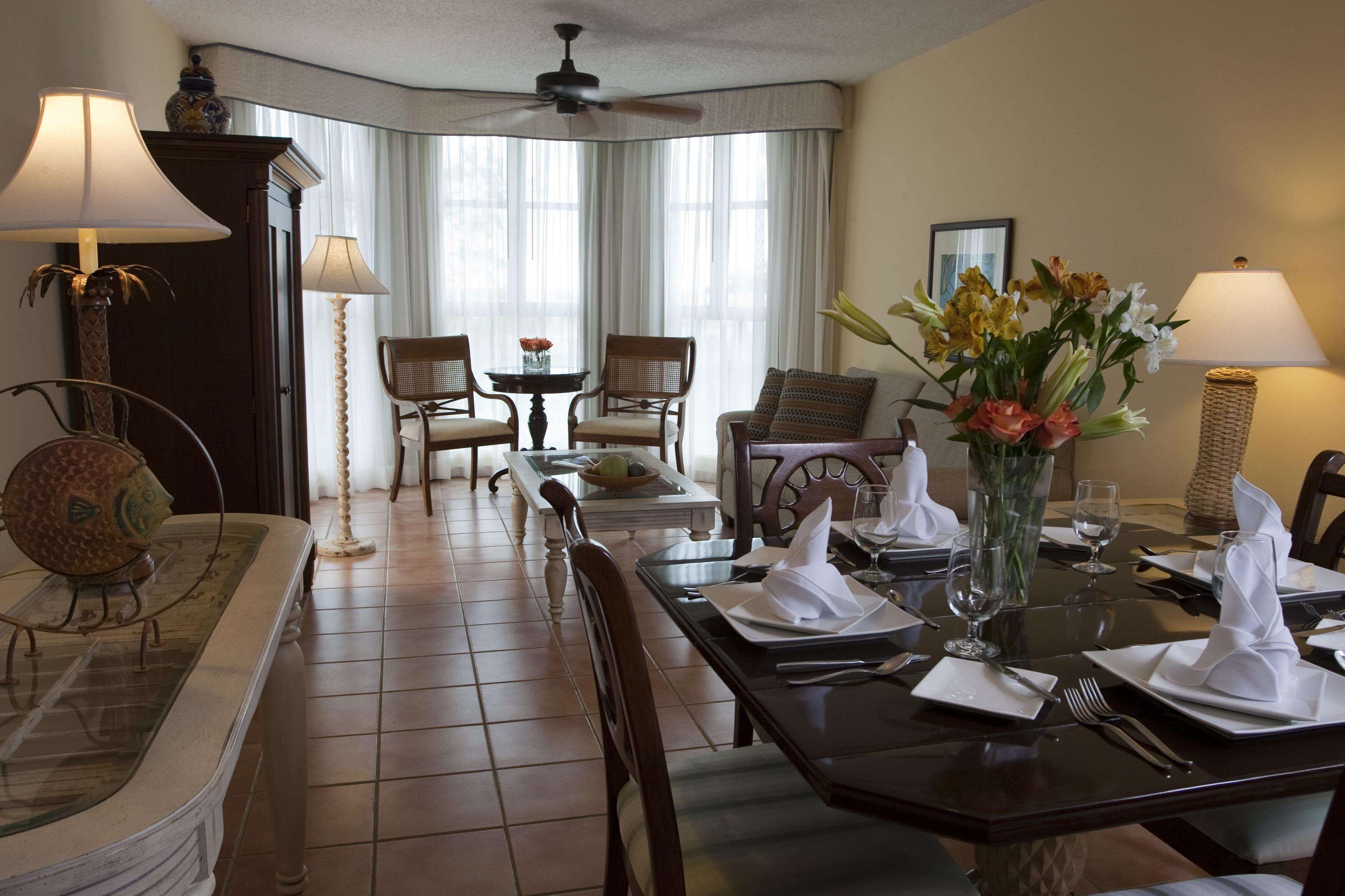 Un comedor con una mesa preparada para cuatro personas, sillas, un florero con flores y un ventilador de techo.