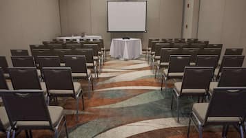 A conference room with rows of chairs facing a white projection screen, a small table with a projector, and a patterned carpet.