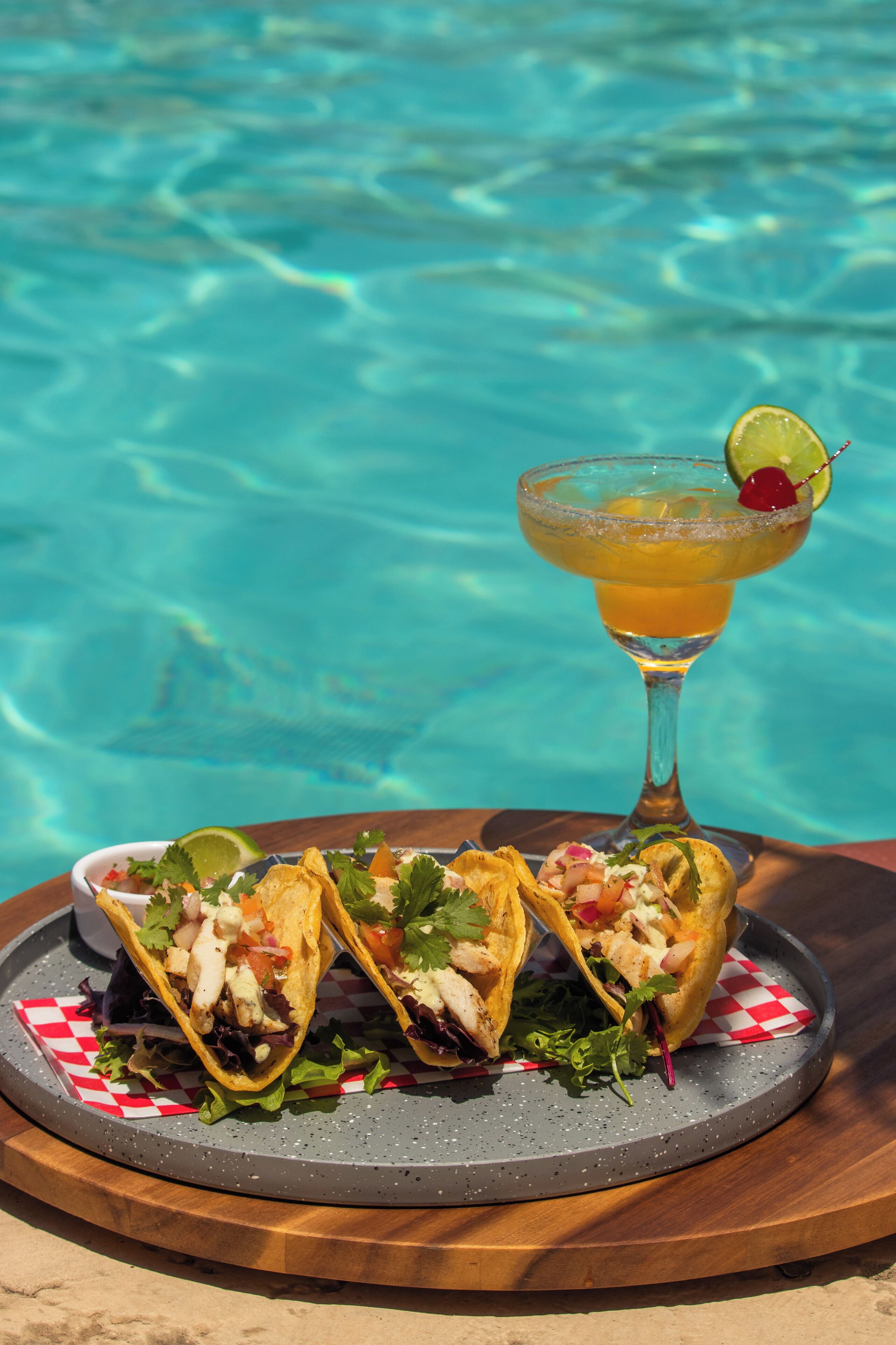 A table with two tacos and a cocktail by a pool.