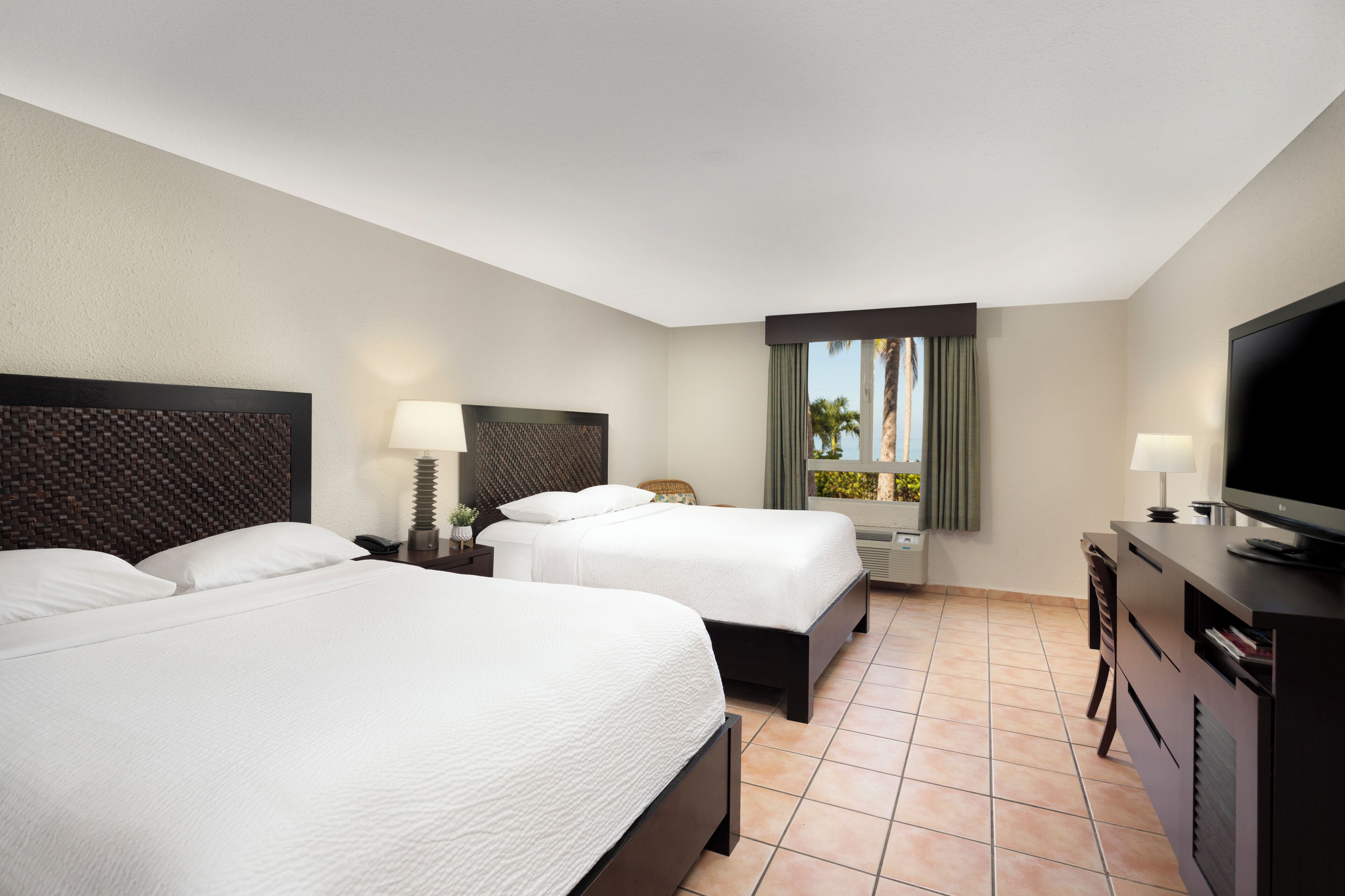A hotel room with two beds, a TV, a desk, and a window with a view of palm trees.