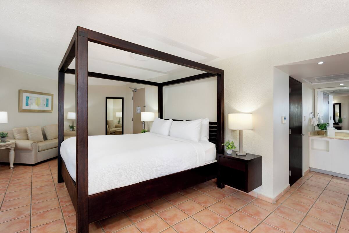 A four-poster bed with white linens, a nightstand with a lamp, a beige sofa, and a bathroom visible through an open door.