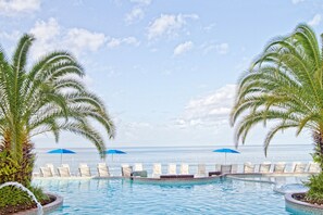 A poolside area with lounge chairs and umbrellas, palm trees, and a view of the ocean.