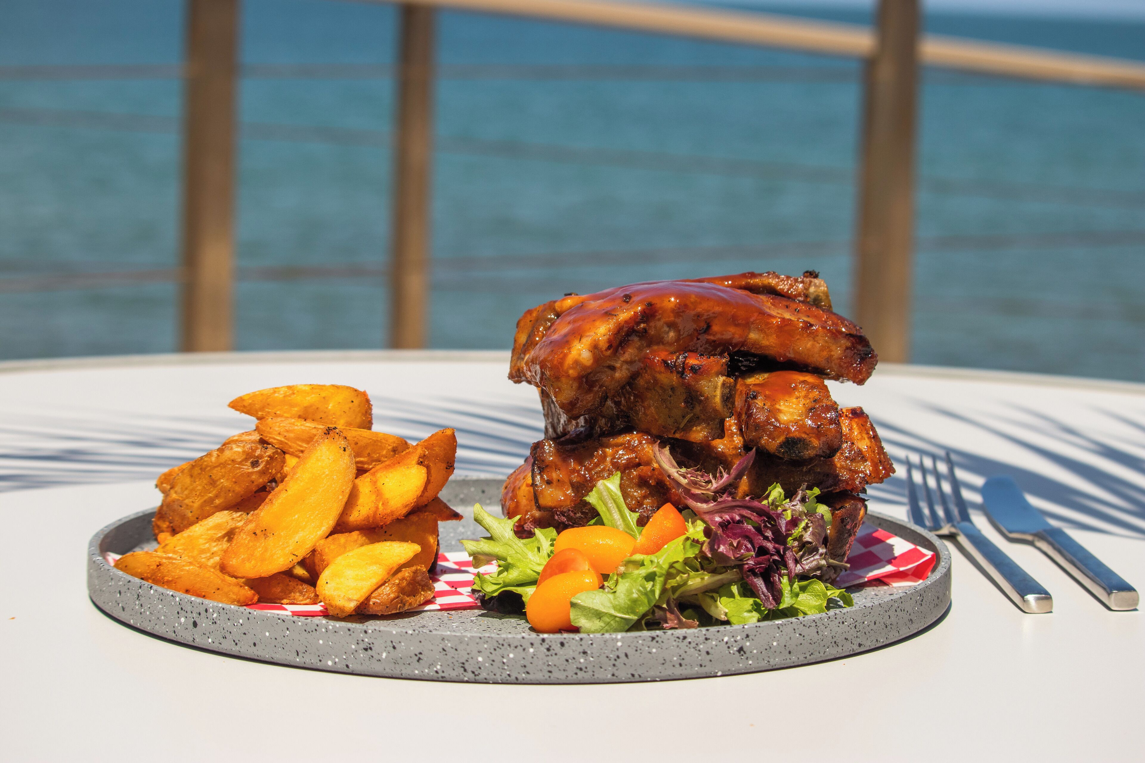 Un piatto di cibo con patate arrosto, verdure a foglia e carne, su un tavolo con vista sul mare.