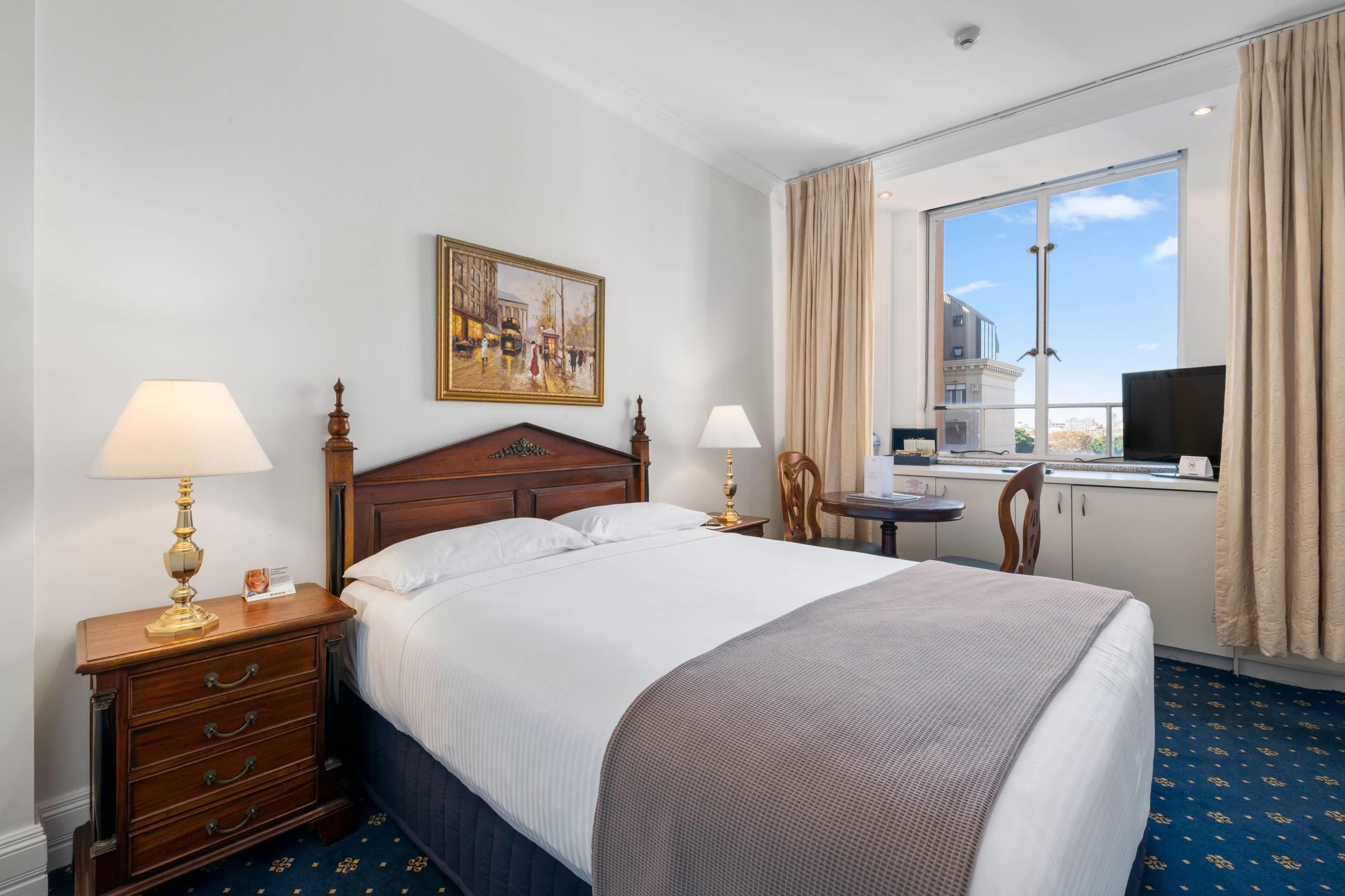 Een hotelkamer met een groot bed, twee nachtkastjes met lampen, een klein bureau met stoel, een televisie en een raam met gordijnen.