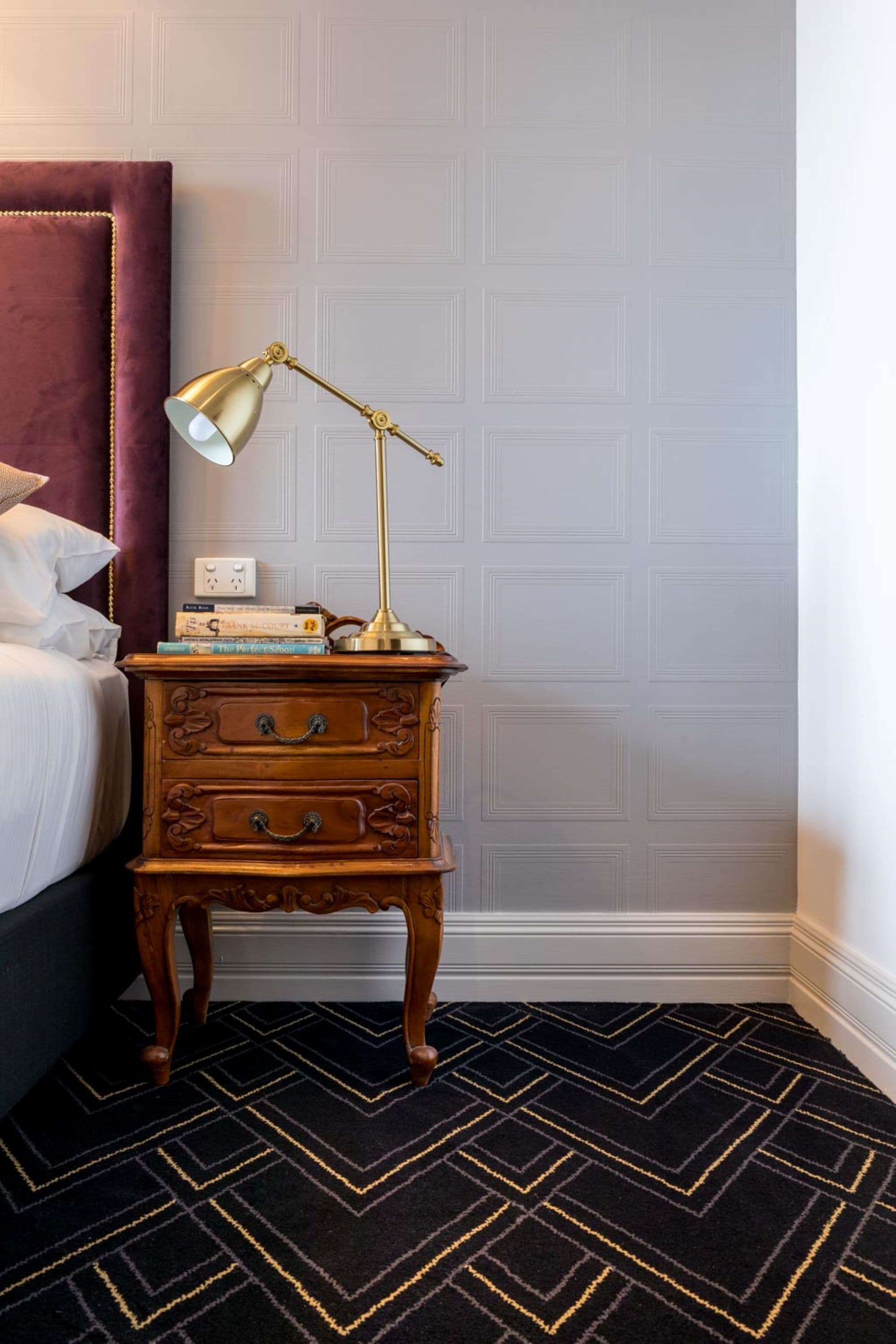 A neatly made bed with a purple headboard, a wooden nightstand with a brass lamp, and a patterned rug.