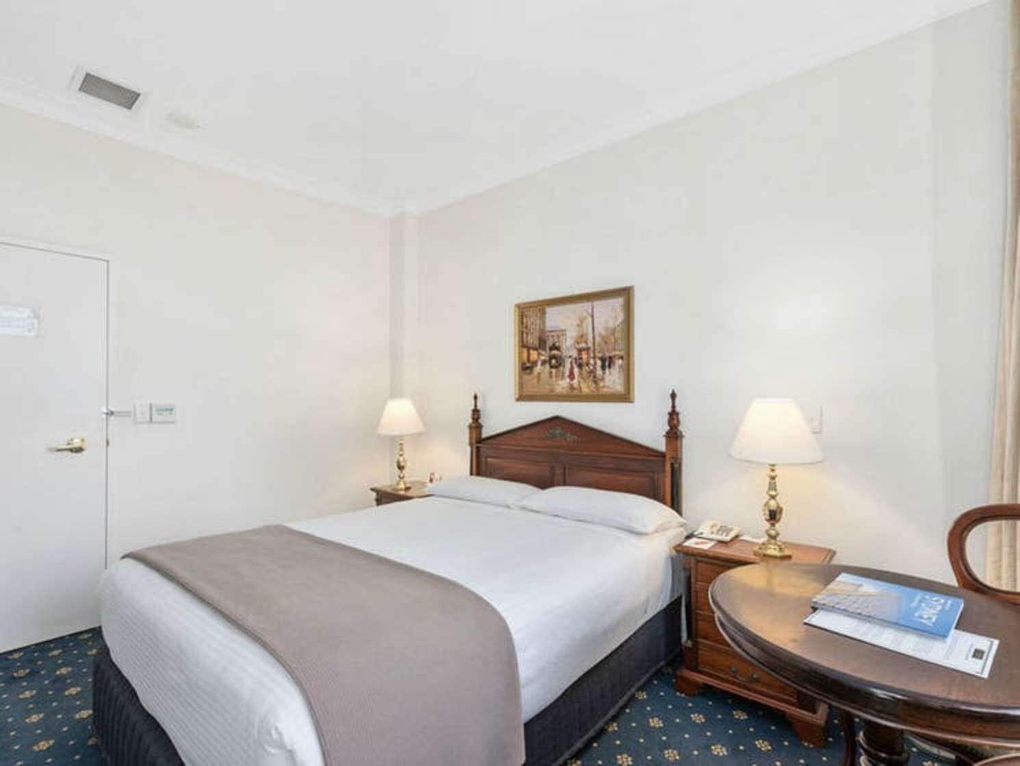 A hotel room with a bed, two bedside tables, a chair, a small table, and a painting on the wall.