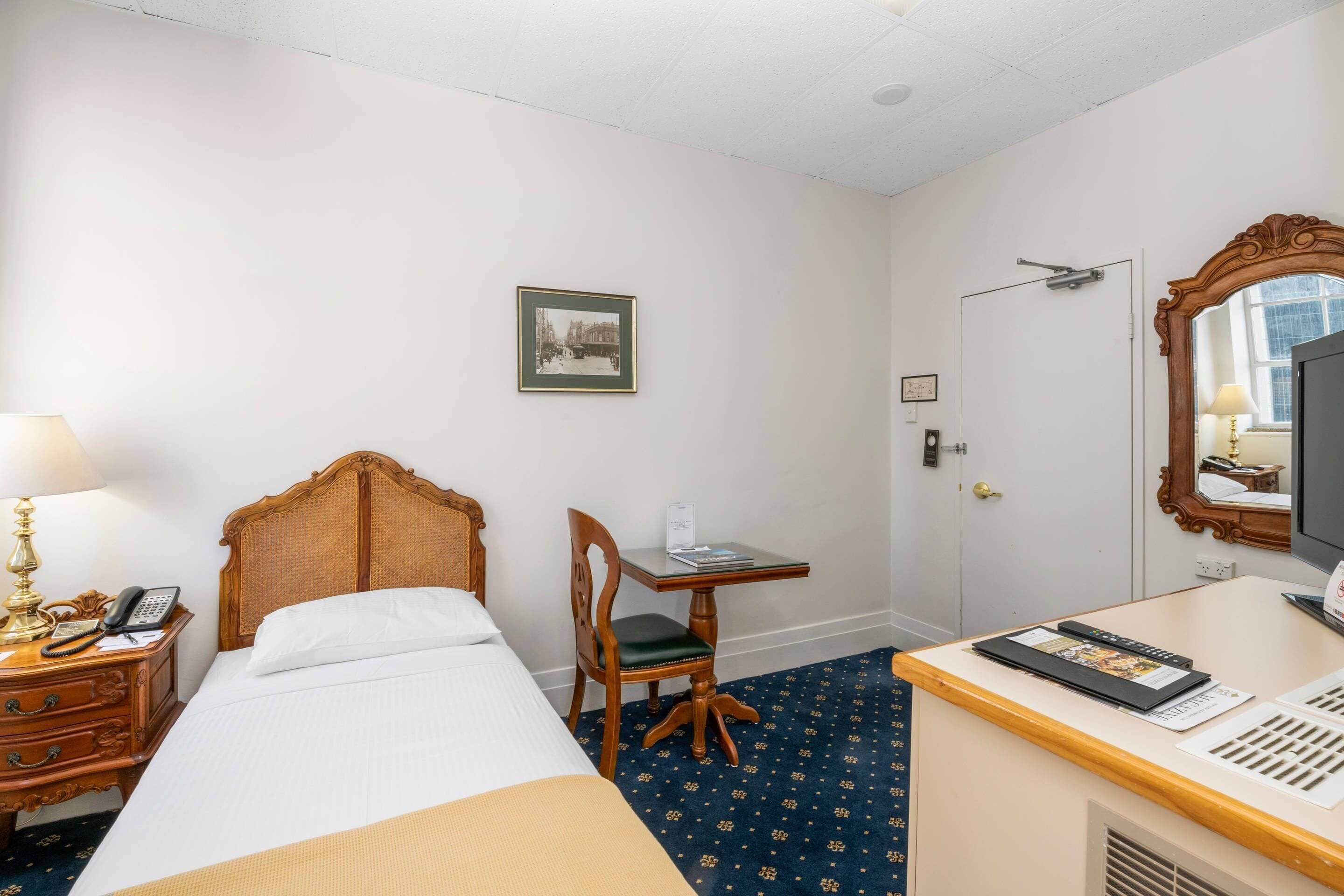 A hotel room with a bed, a desk with a chair, a telephone, a lamp, a mirror, and a picture on the wall.