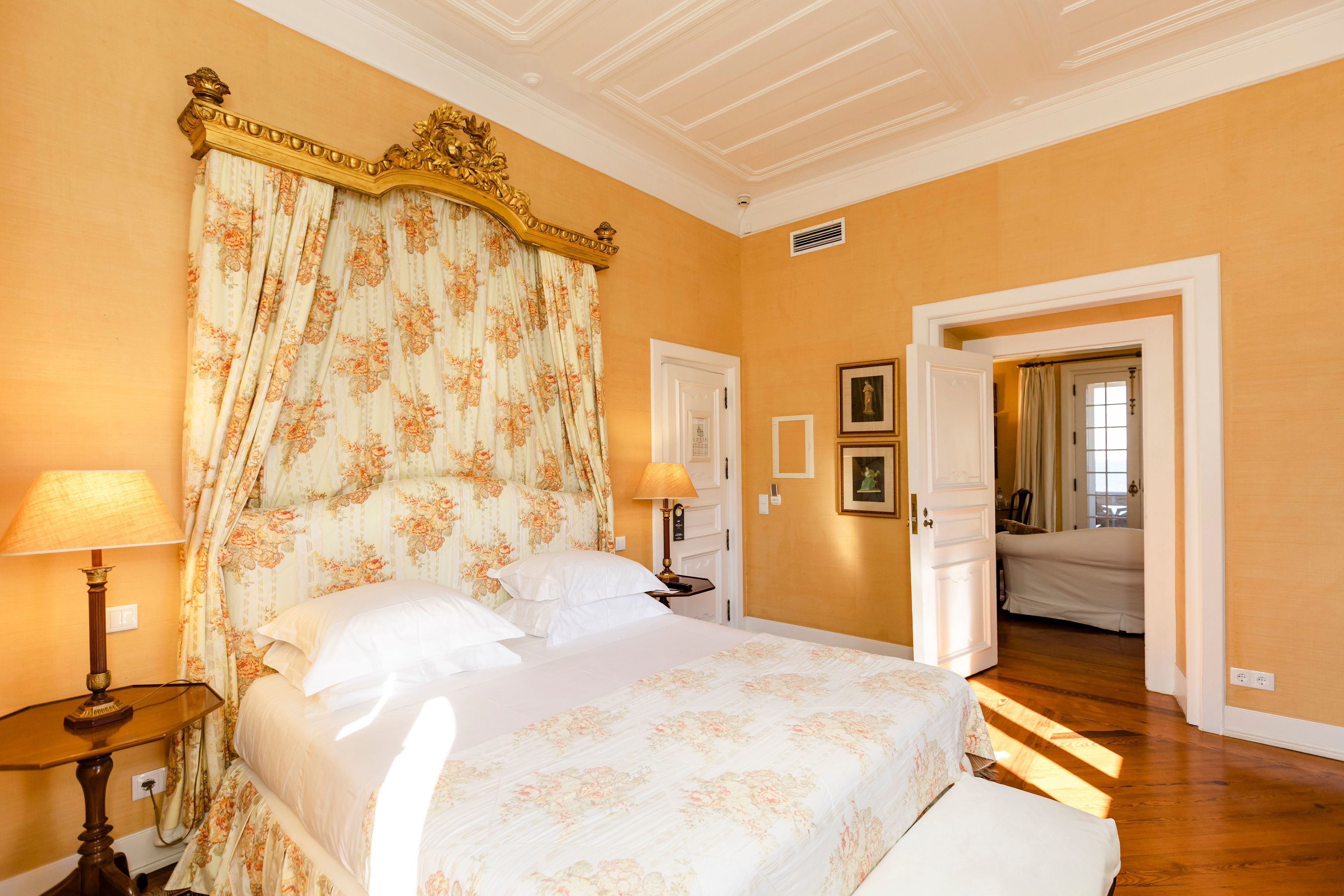 Un dormitorio con una cama grande, cortinas florales, una mesita de noche con lámpara y una puerta que da a otra habitación.