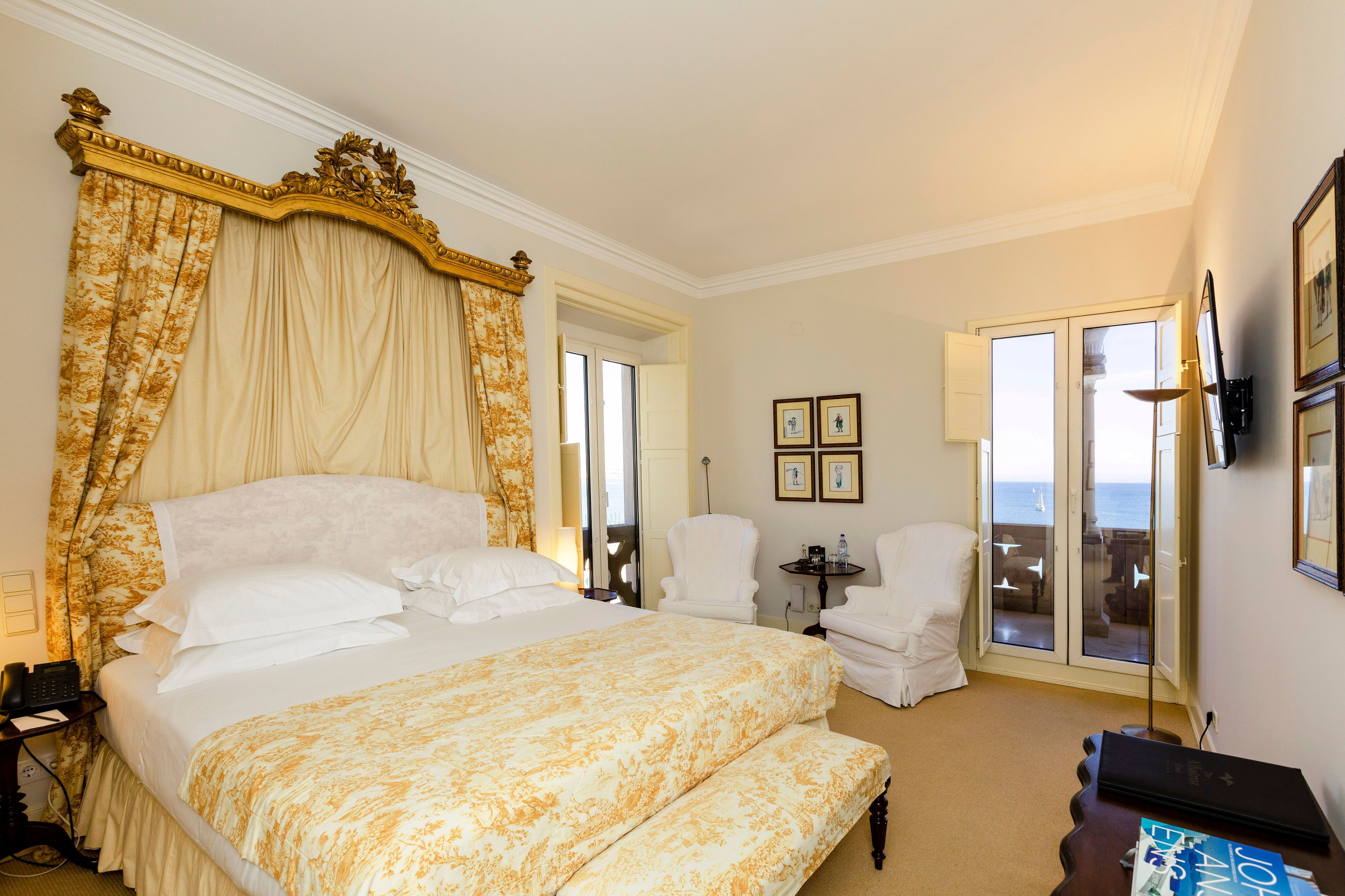 A spacious bedroom with a large bed, a canopy, two white armchairs, a small table, and a view of the sea through a glass door.