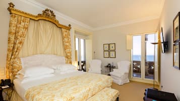 Een ruime slaapkamer met een groot bed, een hemelbed, twee witte fauteuils, een kleine tafel en uitzicht op zee door een glazen deur.