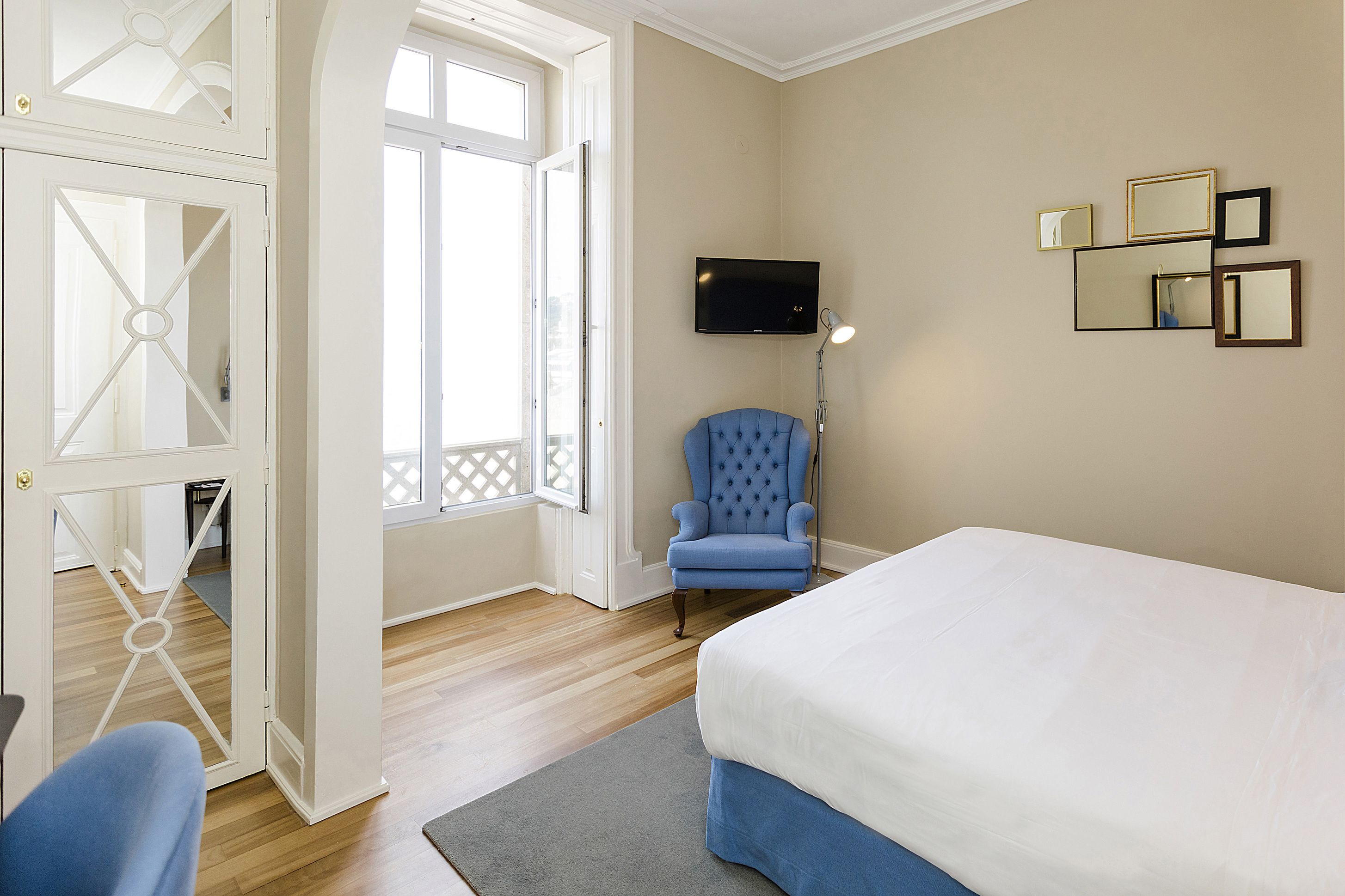 A bedroom with a bed, a blue armchair, a TV, and a door leading to another room.
