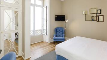 A bedroom with a bed, a blue armchair, a TV, and a door leading to another room.