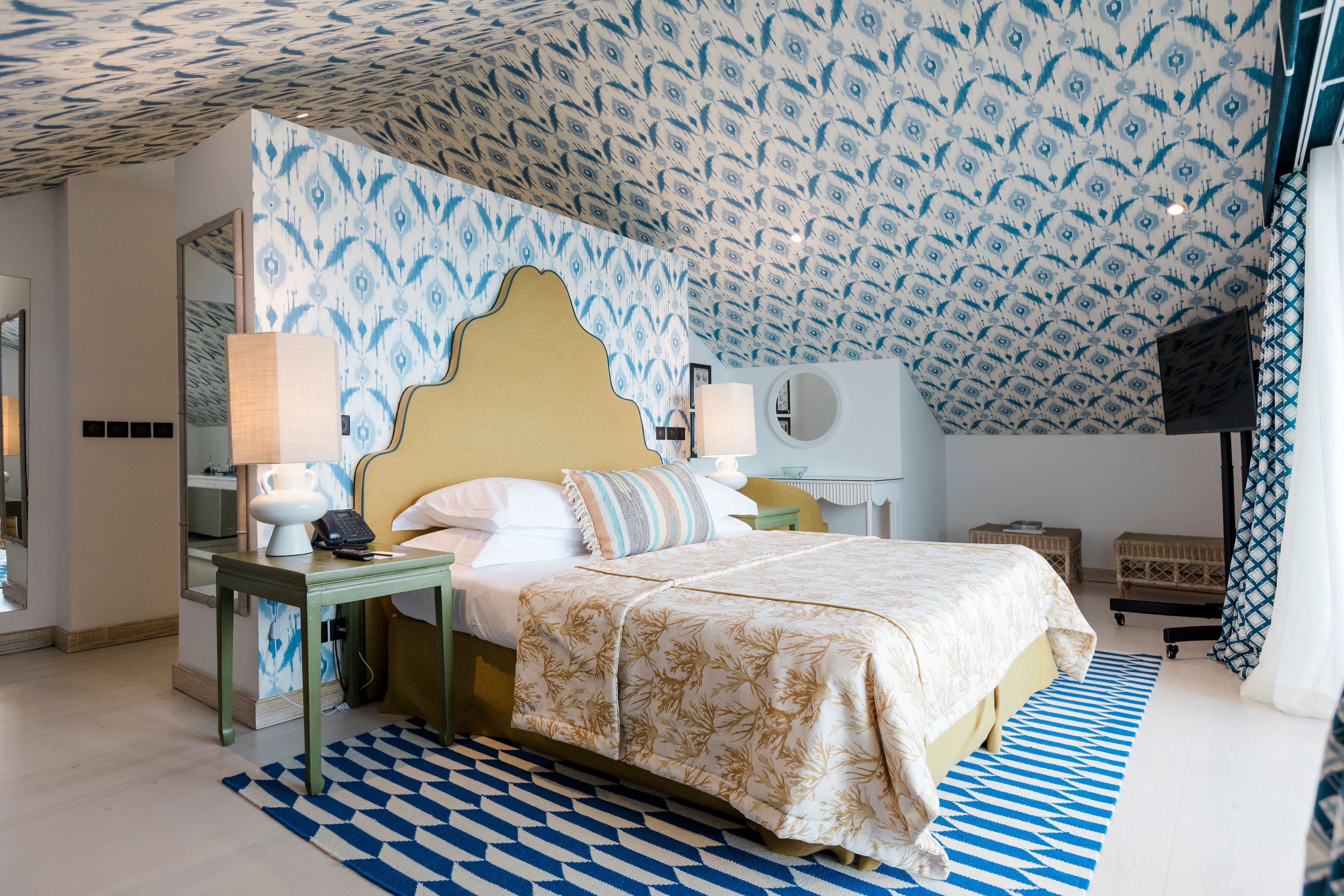 A hotel room with a patterned ceiling, a bed with a floral bedspread, a bedside table with a lamp, and a small round mirror.