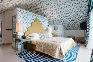 Habitación de hotel con techo estampado, cama con colcha floral, mesita de noche con lámpara y un espejo redondo pequeño.