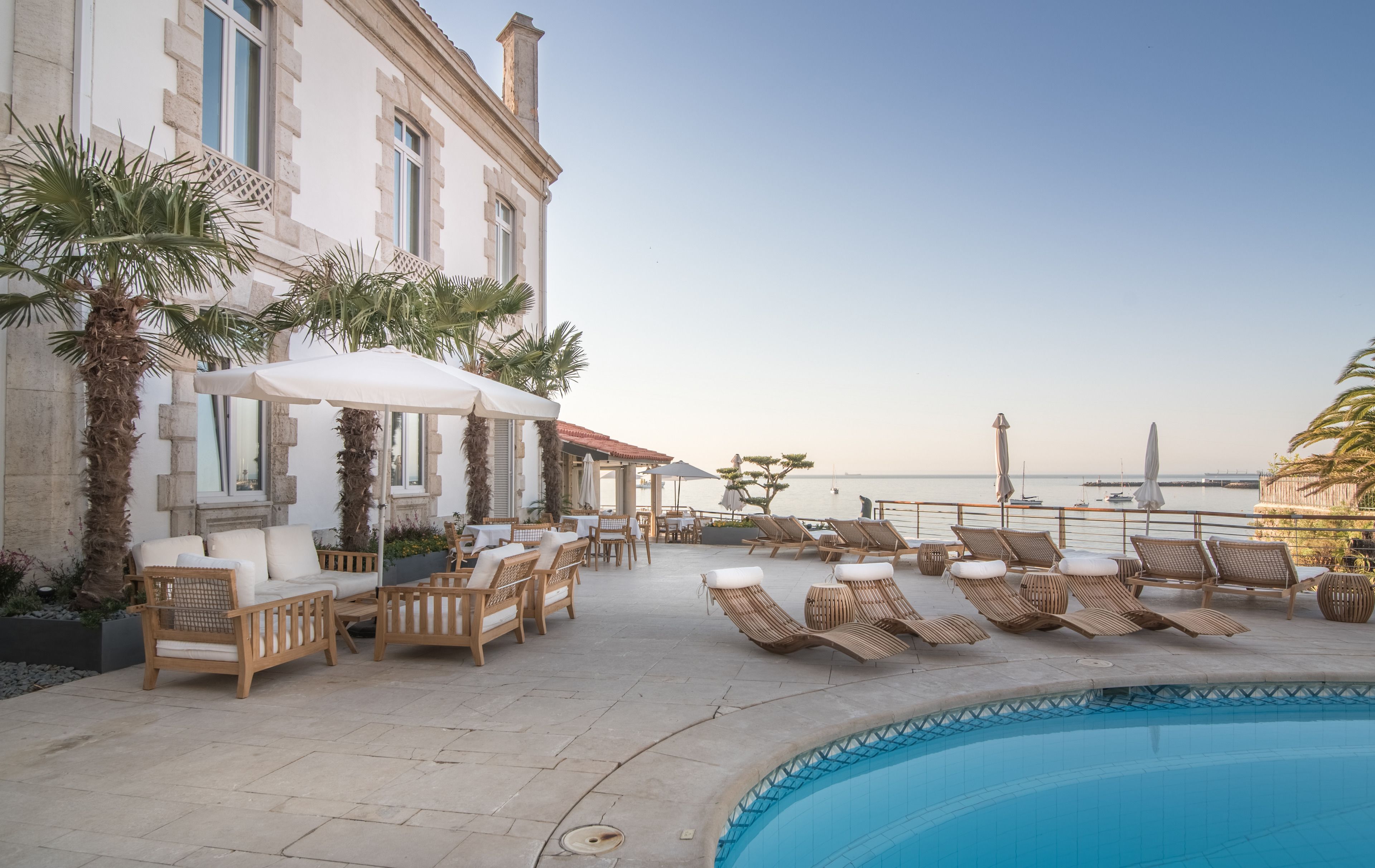A poolside area with lounge chairs and umbrellas, a building with stone facade, and a view of the sea.